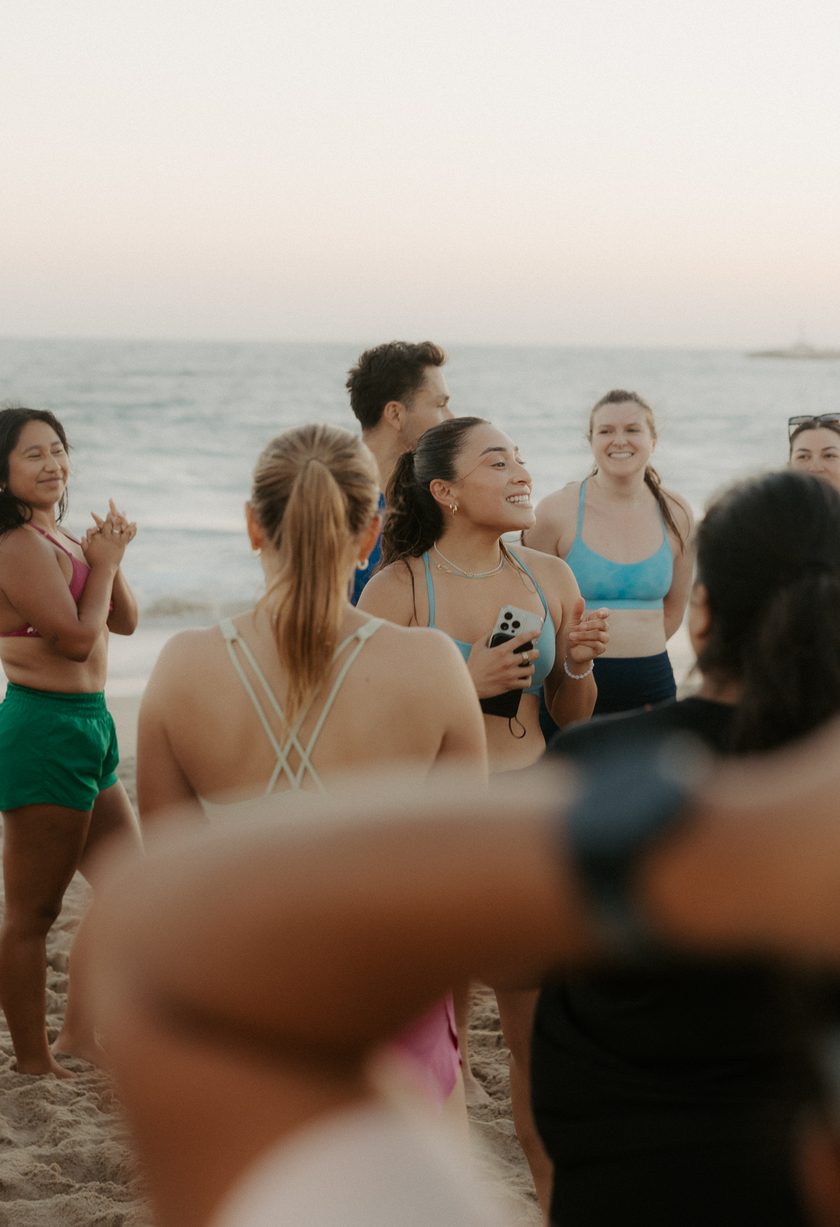 A circle of women laughing and clapping on the beach at sunset